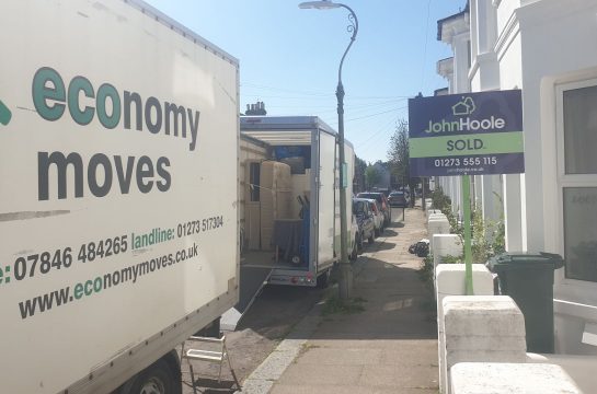 A house with a sold sign outside of it. 2 economy moves vans loading moving boxes into the back of the vans.